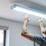A photograph of a lighting contractor installing a 4-ft fluorescent light fixture in a commercial space