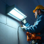 A photograph of a lighting contractor installing a wall pack light on a commercial building exterior at dusk