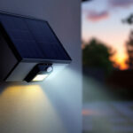 A photograph of a solar-powered flood light with a motion sensor installed in a landscaped outdoor setting at dusk