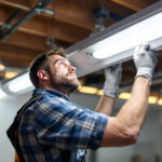 A photograph of a lighting contractor installing or adjusting a 4 ft fluorescent light fixture in a commercial or residential setting