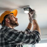 A photograph of a professional lighting contractor installing or inspecting an electrical junction box light fixture in a residential setting