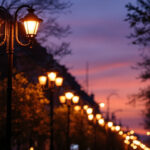 A photograph of a well-lit urban street scene featuring various lamp poles at dusk