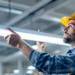 A photograph of a professional lighting contractor installing or inspecting a modern fluorescent lighting fixture in a commercial setting