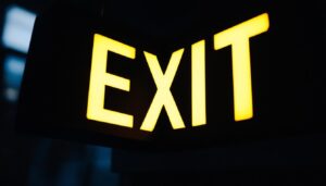 A photograph of a close-up of a black exit sign illuminated in a dimly lit environment