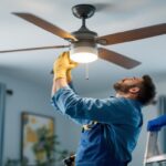 A photograph of a lighting contractor carefully replacing a ceiling fan light fixture in a well-lit room